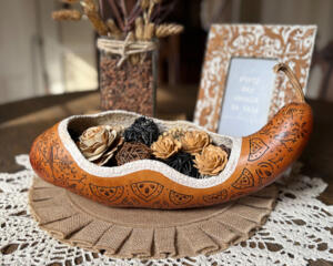 A rustic table centerpiece featuring a real gourd carved into a long decorative bowl filled with wood flowers. It sits on a burlap cloth.